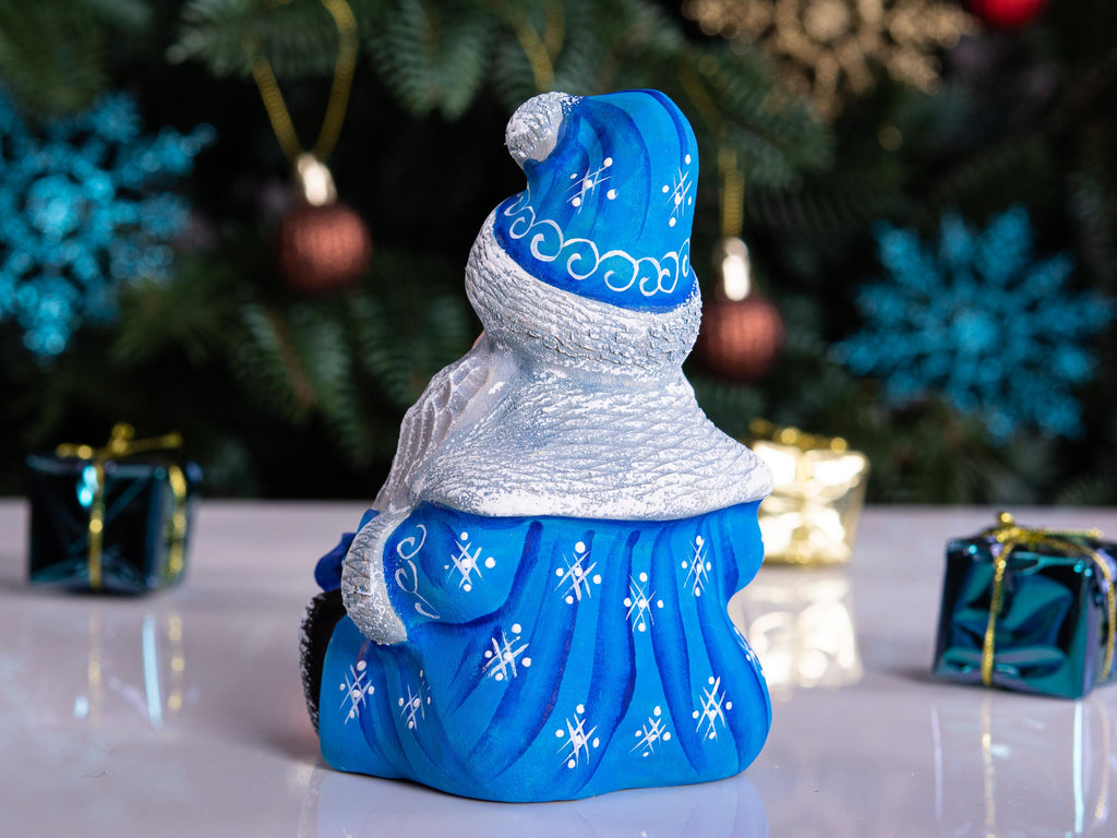 A blue and white Santa Claus Christmas ornament shown from the back, featuring a textured blue hat and sleigh, set against a blurred Christmas tree and gift background.