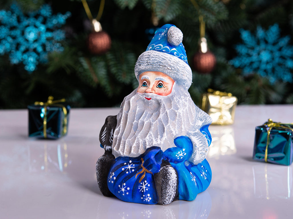 A festive Santa Claus figurine sitting on a reflective white surface, with a blurred Christmas tree and blue snowflake decorations in the background. Two small wrapped gift boxes are placed on either side.