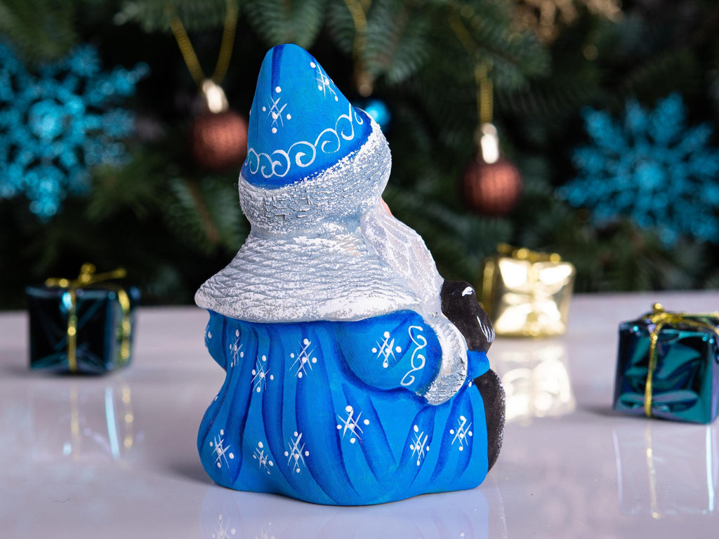 A blue and silver Christmas gnome figurine with a white beard, posed on a reflective white surface with a blurred Christmas tree and gift boxes in the background.