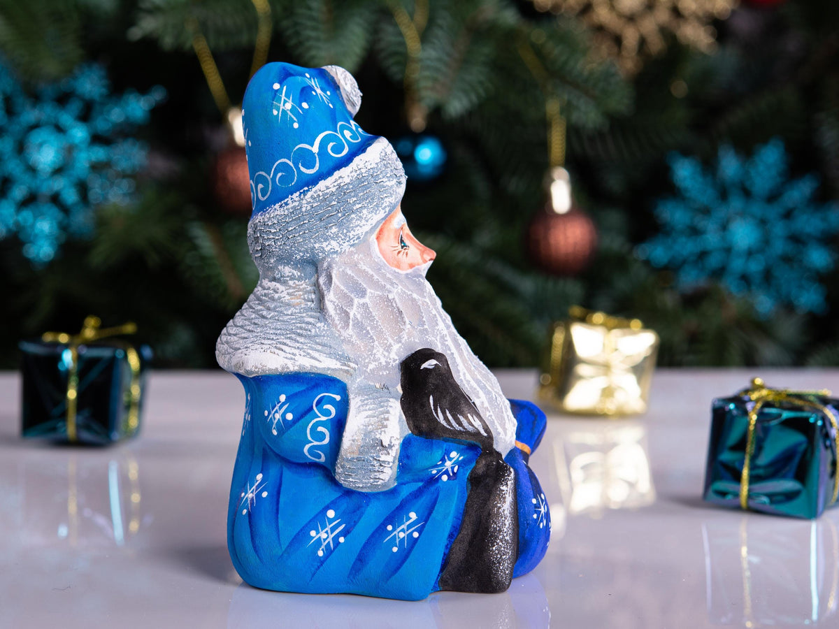 A hand-painted Christmas ornament depicting Santa Claus sitting in a blue armchair with a black cat, set against a blurred Christmas tree background with gift boxes on a reflective white surface.