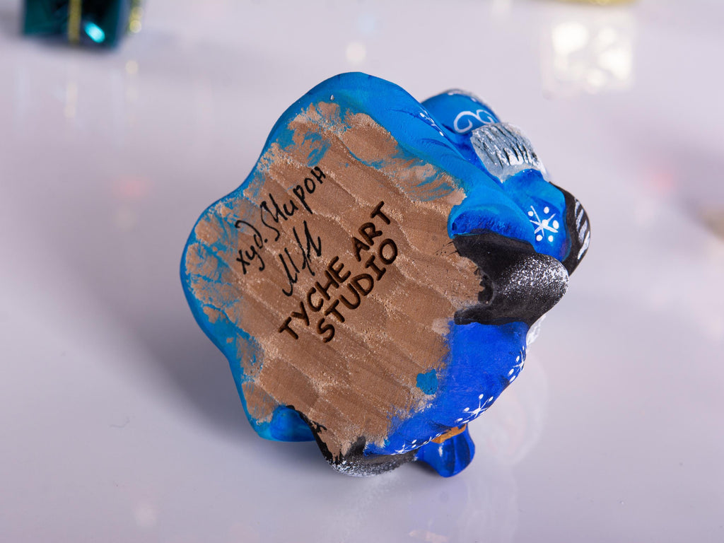 A hand-painted blue and brown ceramic ornament shaped like a sheep, signed by artist Judy Blaker and featuring 'TycHE Art Studio' on the side, photographed on a light reflective surface.