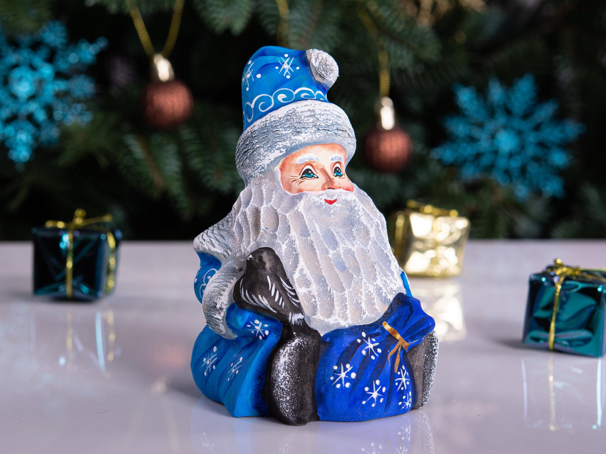 A Santa Claus figurine wearing a blue and white outfit with a white beard, seated on a reflective white surface with blurred Christmas decorations in the background.
