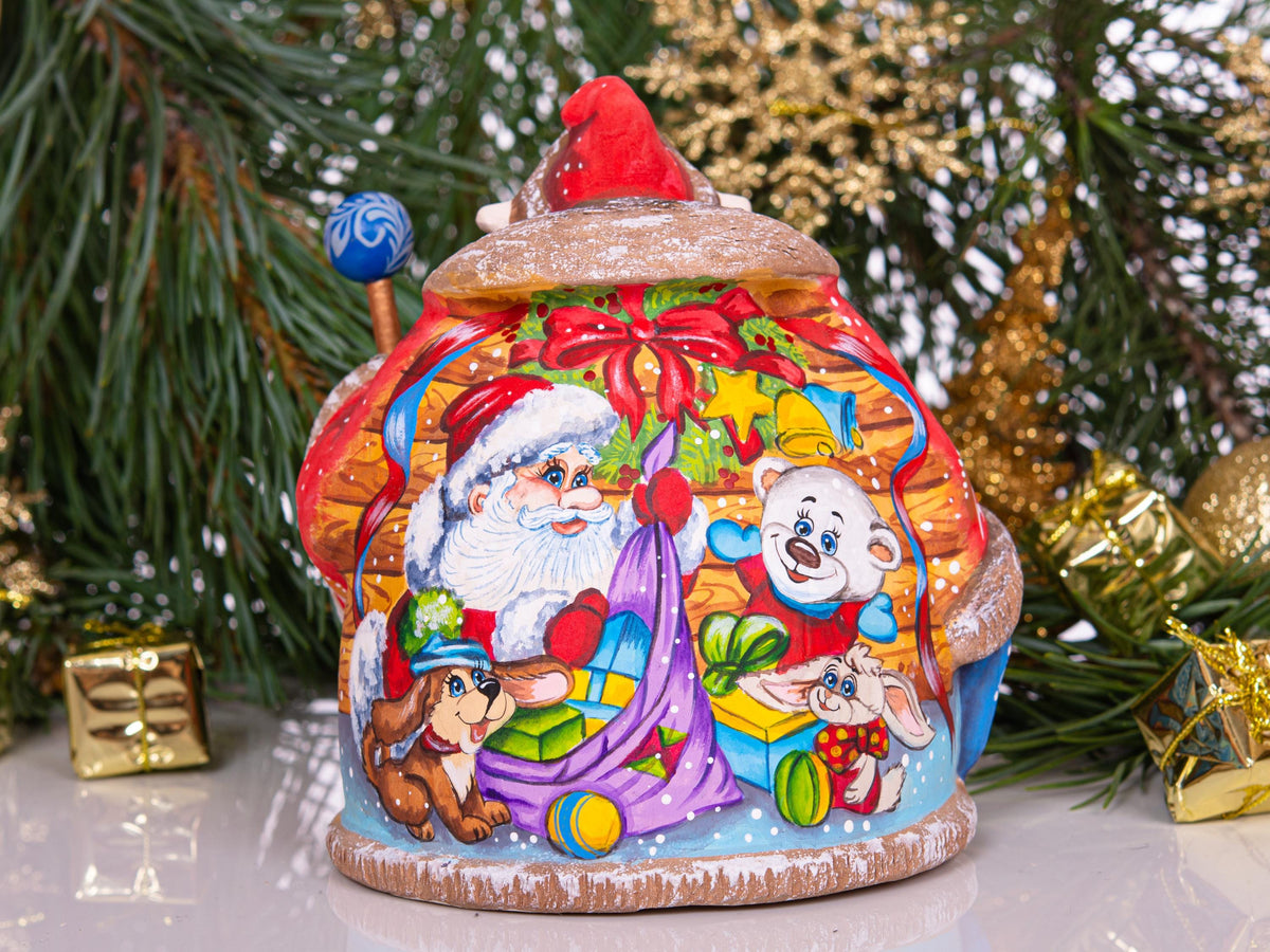 A hand-painted Christmas gingerbread house ornament featuring Santa Claus, a white bear, a rabbit, and a dachshund dog carrying a gift sack, set against a festive background of pine branches and gold tinsel.