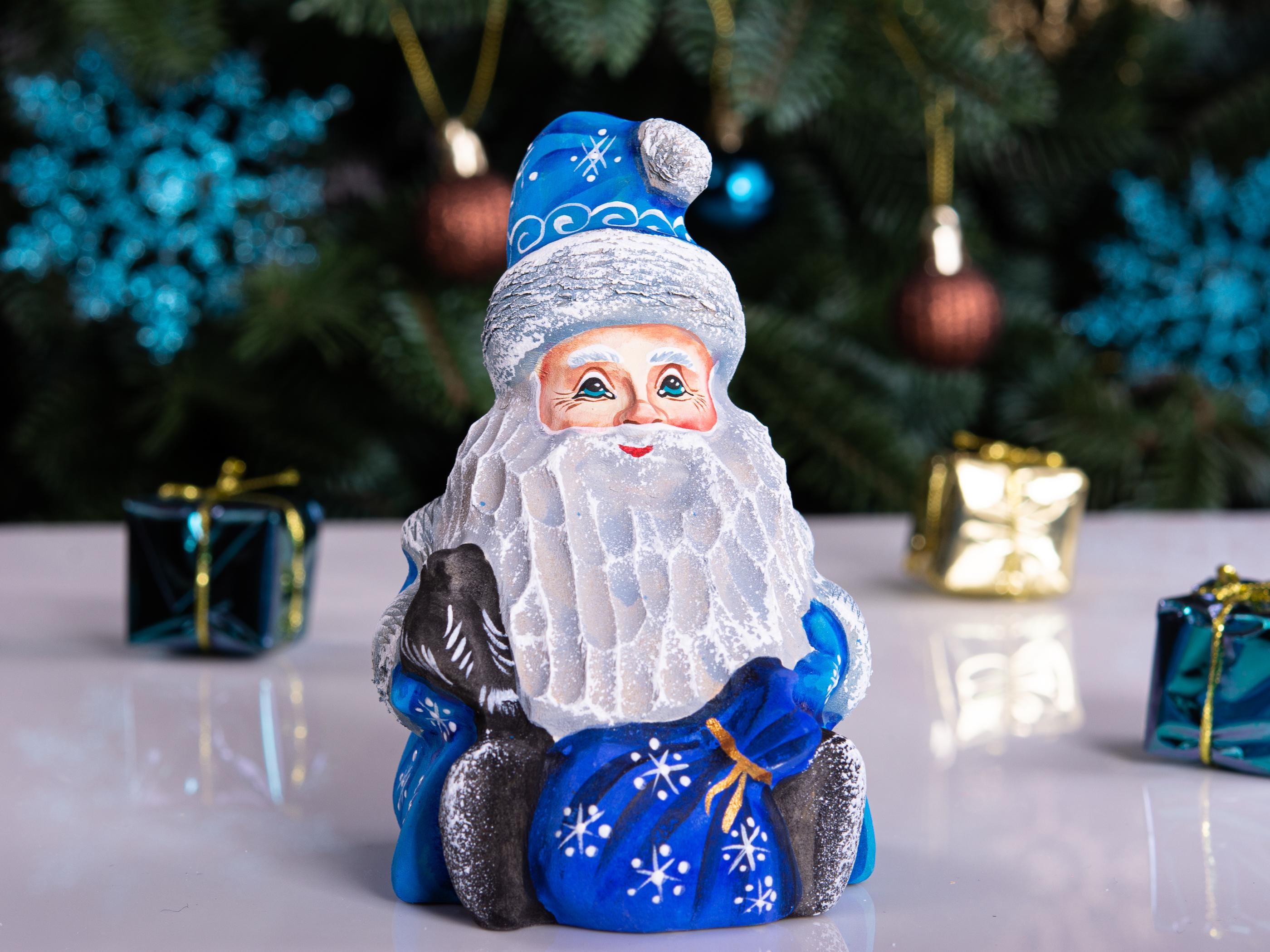 A hand-painted glass Christmas ornament depicting Santa Claus with a white beard, blue hat and coat, sitting on a sleigh, displayed on a reflective white surface with a blurred Christmas tree in the background.