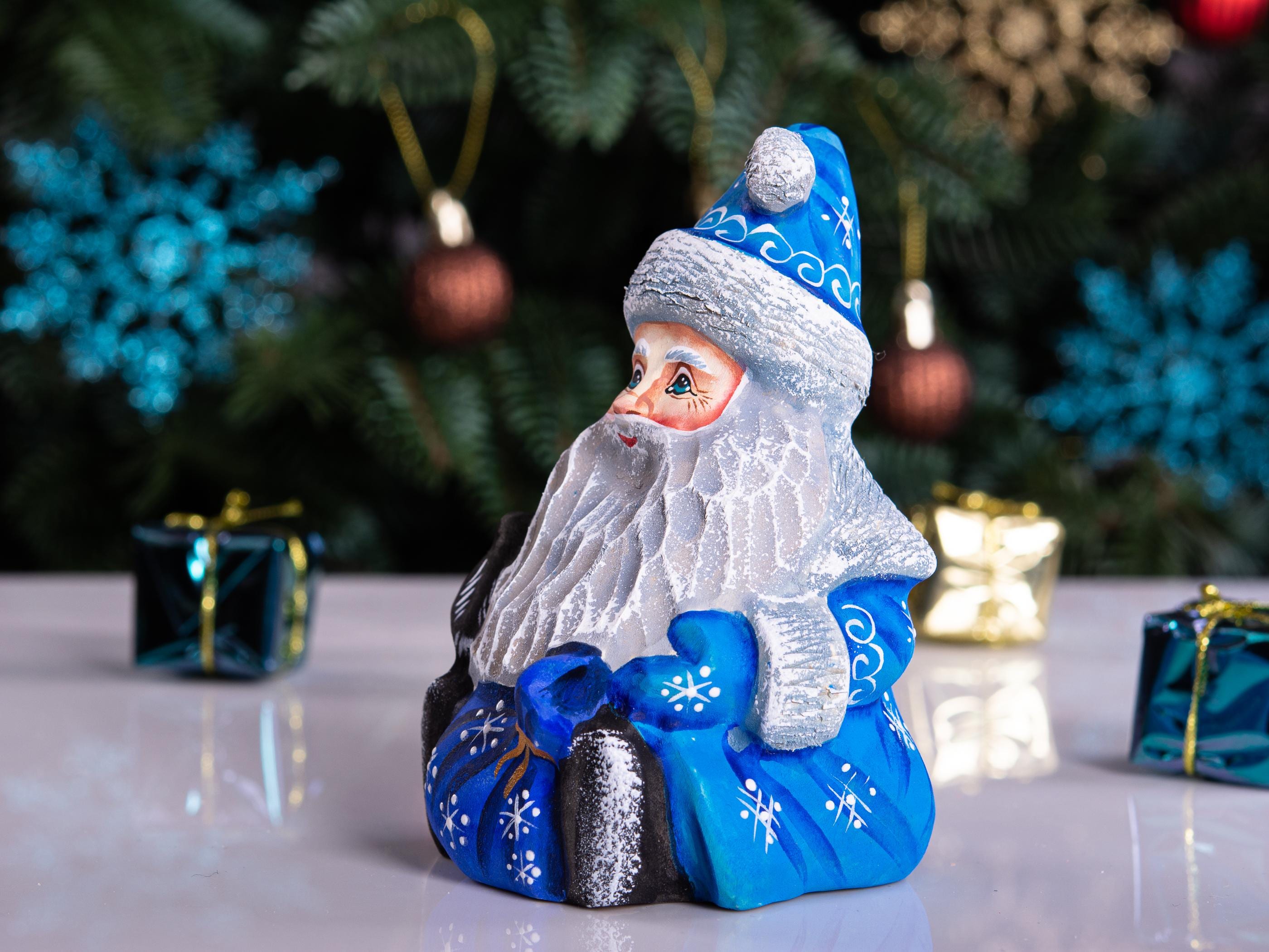 A Santa Claus figurine with a white beard, blue hat and coat decorated with white snowflake patterns, sitting on a reflective white surface with a blurred Christmas tree and gift boxes in the background.