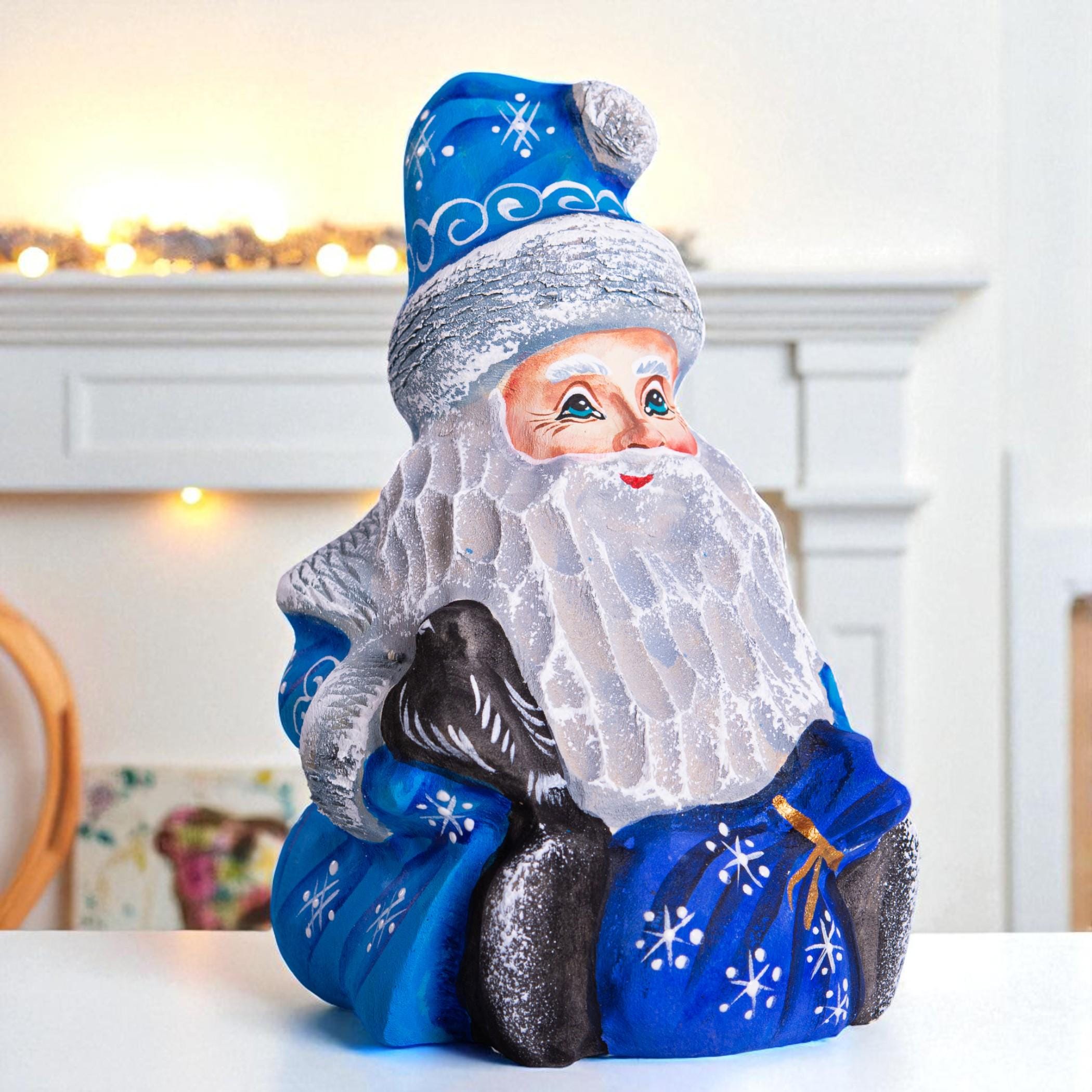 A Santa Claus figurine or decorative bust with a blue hat and coat, white beard, and black boots, shown sitting on a white surface in front of a softly blurred indoor background.