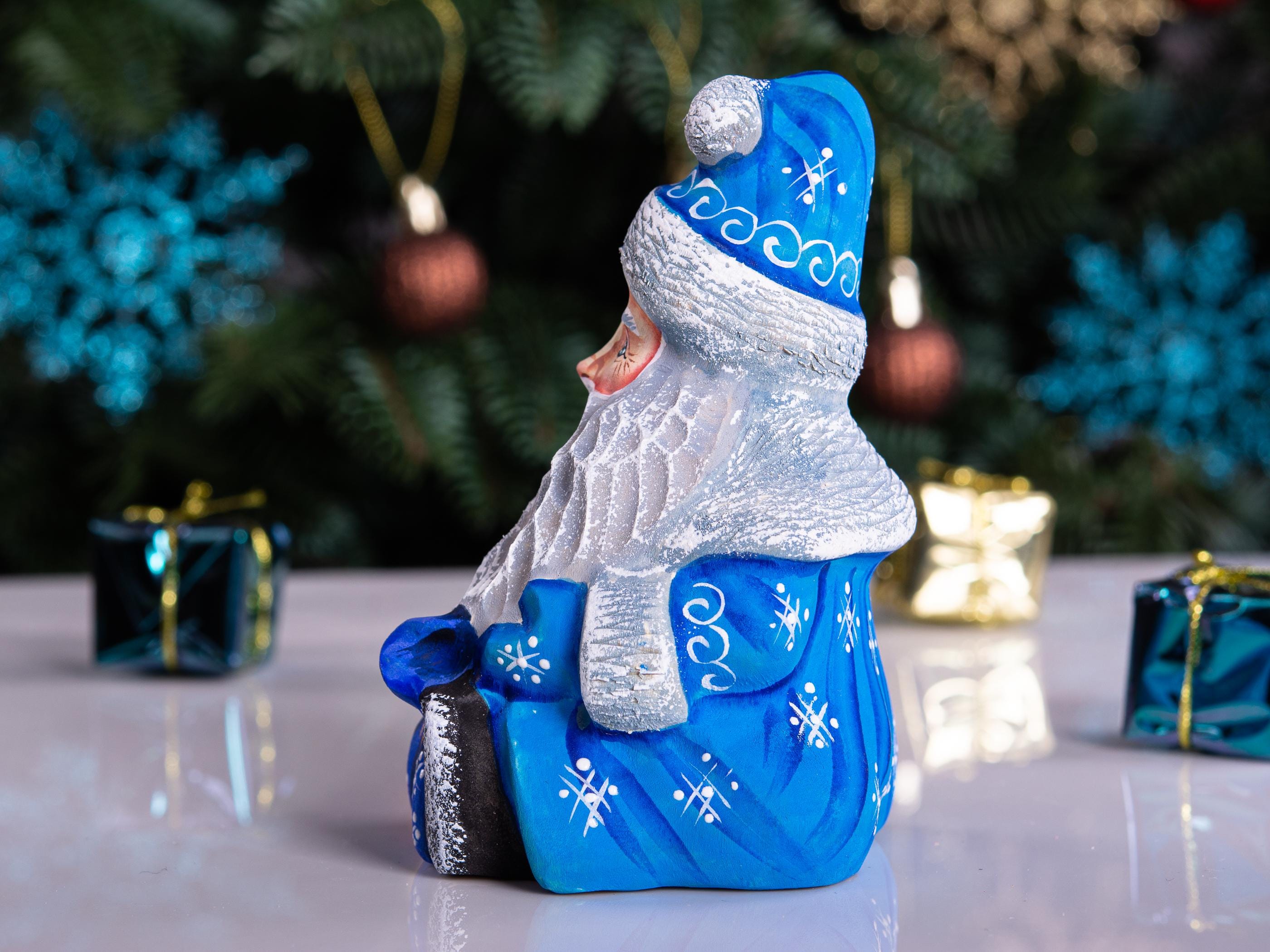 A blue and silver Santa Claus figurine sitting on a reflective white surface, with a blurred Christmas tree and gift boxes in the background.